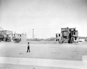 a black and white photograph of a boy walking down a mostly demolished residential street. Two clusters of derelict homes still stand.